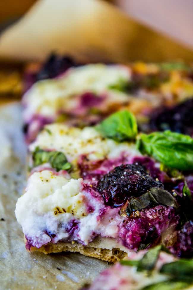 Hands spreading ricotta on pizza dough with fresh blackberries and basil leaves arranged nearby on marble counter