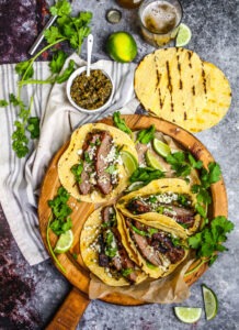 Perfectly grilled carne asada tacos garnished with fresh cilantro, diced onions, and lime wedges on a wooden serving board