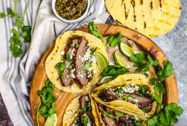 Perfectly grilled carne asada tacos garnished with fresh cilantro, diced onions, and lime wedges on a wooden serving board