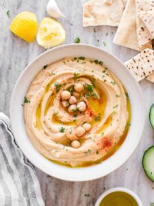Smooth and creamy homemade hummus in a white bowl, drizzled with golden olive oil and sprinkled with paprika, surrounded by fresh vegetables and warm pita bread