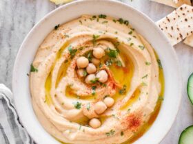 Smooth and creamy homemade hummus in a white bowl, drizzled with golden olive oil and sprinkled with paprika, surrounded by fresh vegetables and warm pita bread