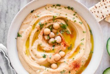 Smooth and creamy homemade hummus in a white bowl, drizzled with golden olive oil and sprinkled with paprika, surrounded by fresh vegetables and warm pita bread