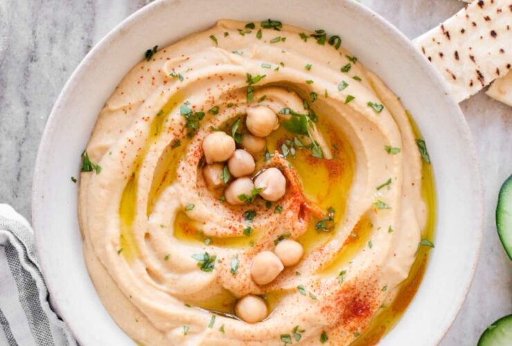 Smooth and creamy homemade hummus in a white bowl, drizzled with golden olive oil and sprinkled with paprika, surrounded by fresh vegetables and warm pita bread