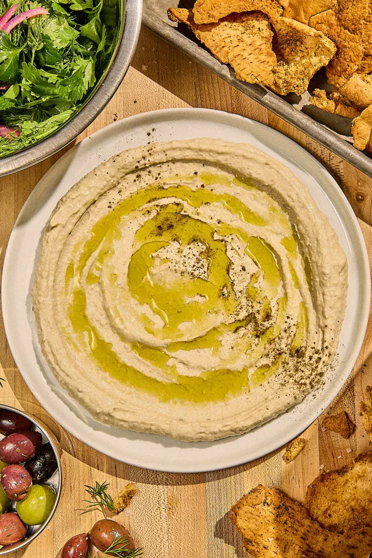 Food processor filled with smooth hummus mixture, showing the creamy texture, with ingredients like tahini jar, lemon halves, and olive oil bottle arranged nearby
