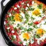 Shakshuka served in cast iron skillet with golden egg yolks and fresh herbs, garnished with crumbled feta and served with crusty bread