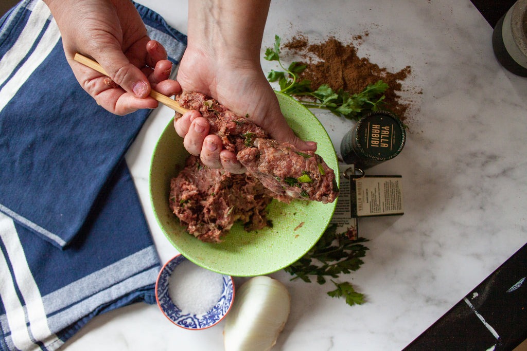 Step-by-step shot showing hands shaping kafta mixture around metal skewers with ingredients laid out nearby