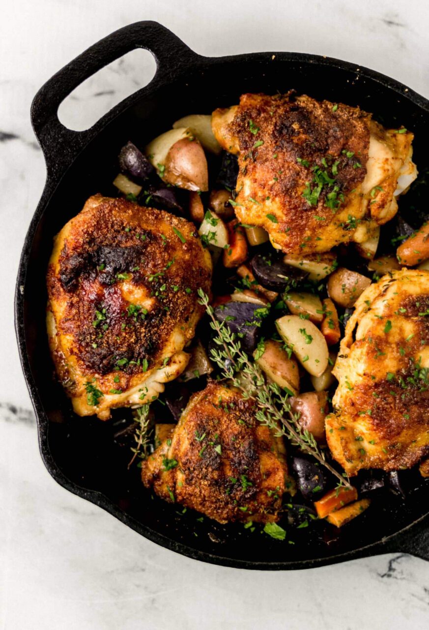 Seasoned chicken thighs in a cast iron skillet with fresh vegetables and herbs arranged on a cutting board nearby
