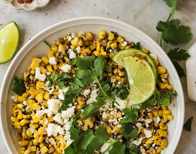 Mexican street corn served in a rustic skillet garnished with fresh cilantro, lime wedges, and crumbled cotija cheese