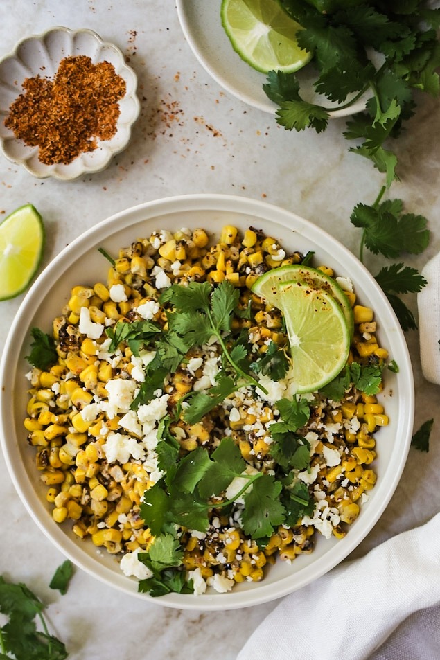 Mexican street corn served in a rustic skillet garnished with fresh cilantro, lime wedges, and crumbled cotija cheese