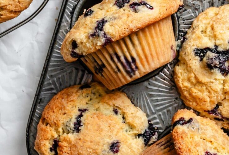 Beautiful golden-brown blueberry muffins with towering tops in a muffin tin, some cooling on a wire rack, with fresh blueberries scattered around