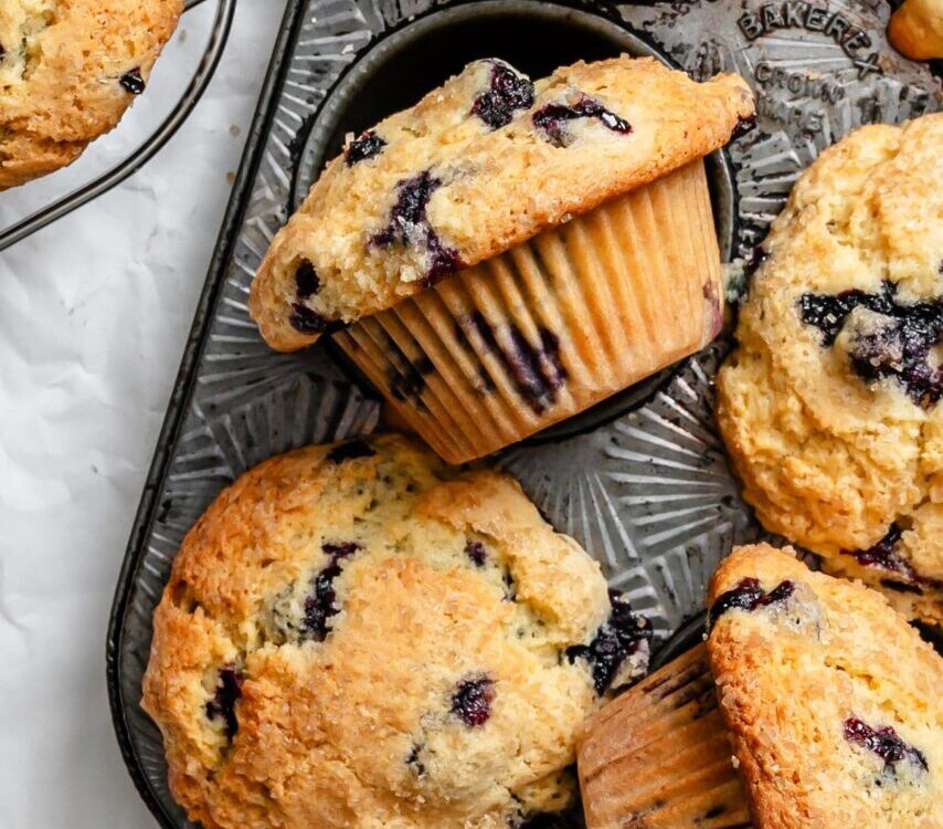 Beautiful golden-brown blueberry muffins with towering tops in a muffin tin, some cooling on a wire rack, with fresh blueberries scattered around