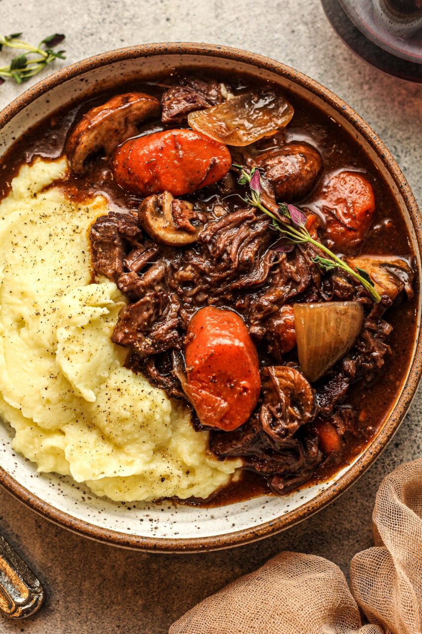 Close-up shot of fork-tender beef with glossy burgundy sauce and herbs, showing the rich texture and color of the finished dish