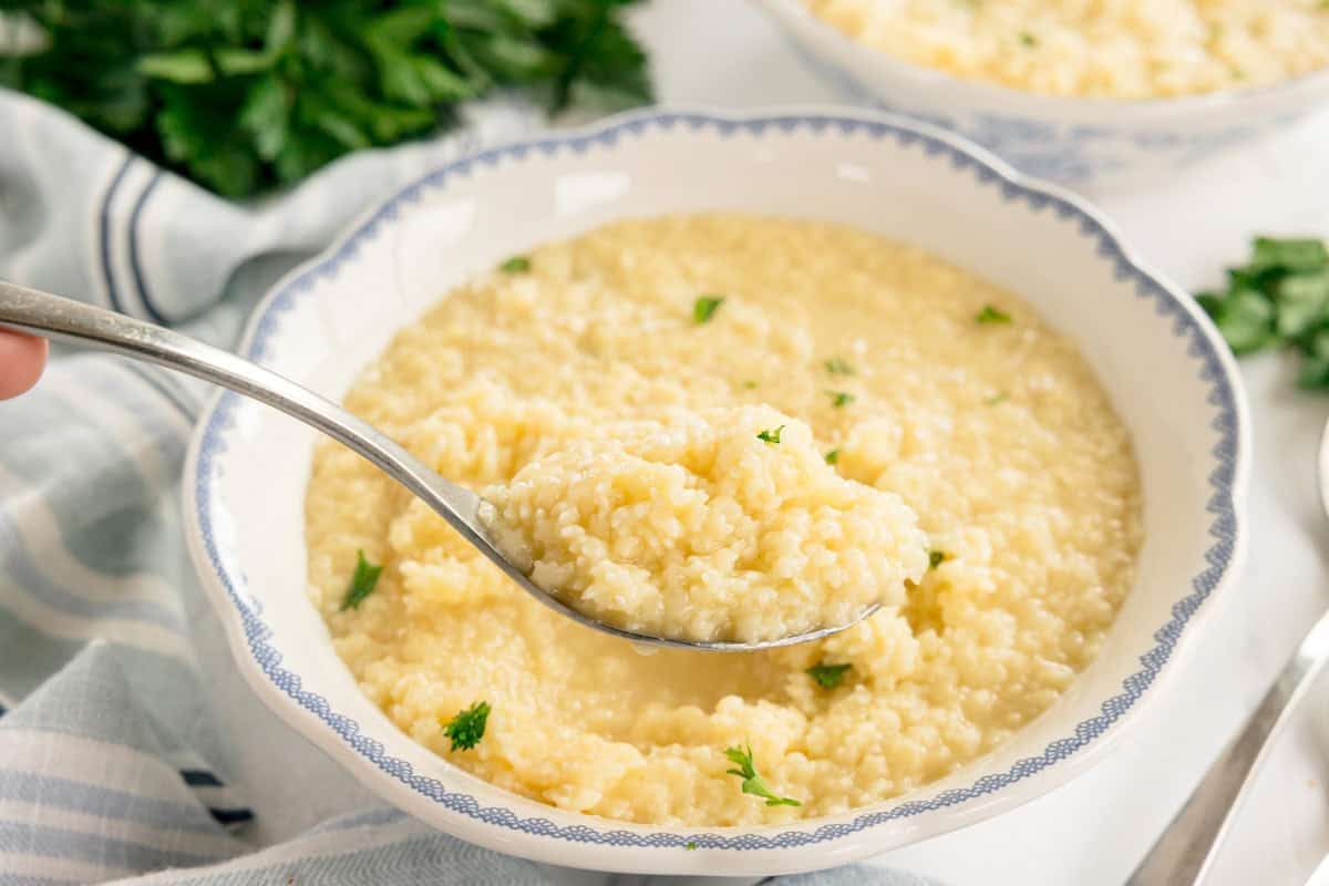 Close-up shot of a spoonful of creamy buttered pastina with melted cheese strings and visible black pepper, showing the perfect glossy texture