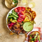 Sliced chicken shawarma on a platter with fresh herbs, pita bread, and colorful vegetables arranged around it