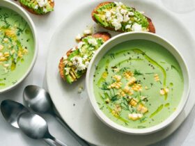Elegant white bowls filled with pale green cold cucumber avocado soup, garnished with fresh herbs and a drizzle of olive oil, photographed from above on a light wooden table with cucumber slices scattered around