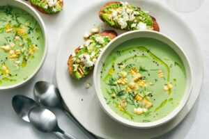 Elegant white bowls filled with pale green cold cucumber avocado soup, garnished with fresh herbs and a drizzle of olive oil, photographed from above on a light wooden table with cucumber slices scattered around