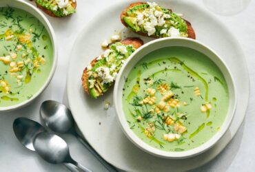 Elegant white bowls filled with pale green cold cucumber avocado soup, garnished with fresh herbs and a drizzle of olive oil, photographed from above on a light wooden table with cucumber slices scattered around