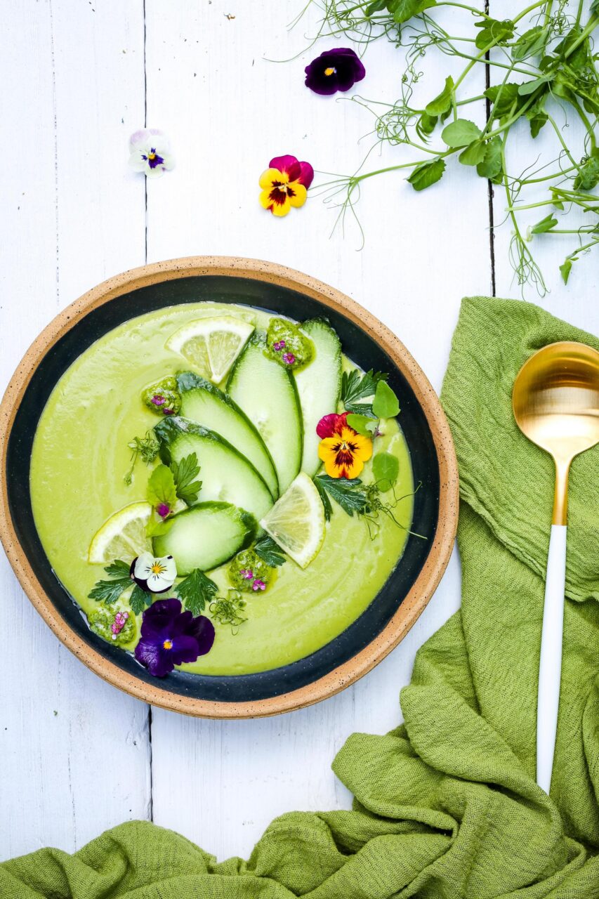 Overhead shot of ingredients laid out on a marble countertop - halved cucumbers, ripe avocados, fresh herbs, yogurt in a bowl, and a high-speed blender in the background