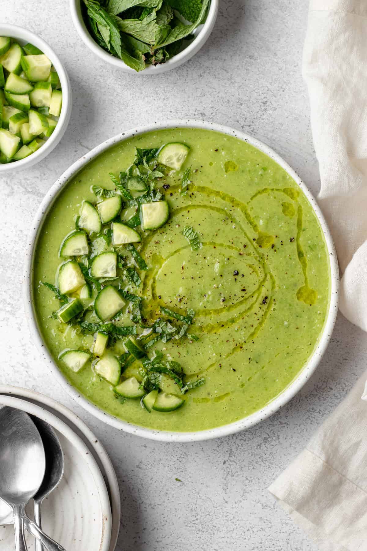 Close-up shot of the cold soup being ladled into a bowl, showing the smooth, creamy texture with visible flecks of fresh herbs, garnished with a cucumber ribbon and served alongside crusty bread