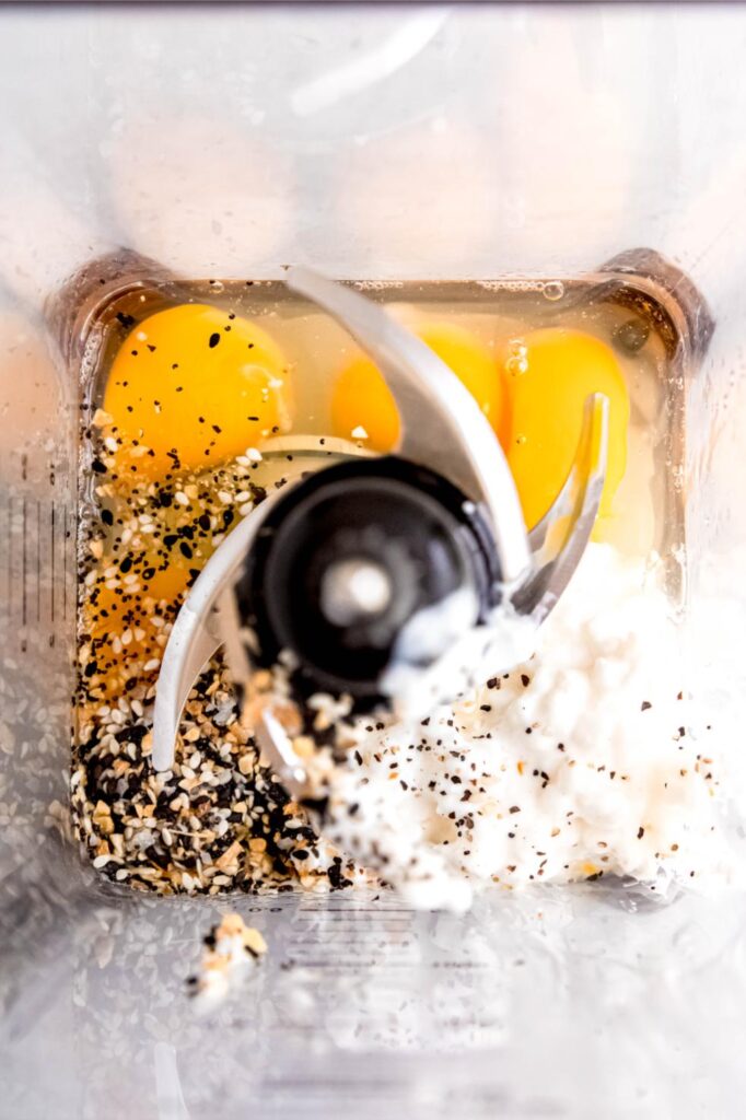 Ingredients for egg bites laid out on a marble counter with cottage cheese being blended until smooth
