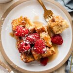 Golden French toast casserole in a white baking dish, served with fresh berries and maple syrup drizzled on top, shot from above on a rustic wooden table with coffee cups visible
