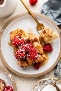 Golden French toast casserole in a white baking dish, served with fresh berries and maple syrup drizzled on top, shot from above on a rustic wooden table with coffee cups visible