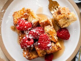 Golden French toast casserole in a white baking dish, served with fresh berries and maple syrup drizzled on top, shot from above on a rustic wooden table with coffee cups visible