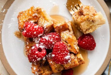 Golden French toast casserole in a white baking dish, served with fresh berries and maple syrup drizzled on top, shot from above on a rustic wooden table with coffee cups visible