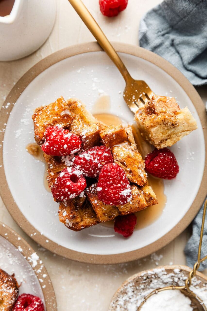 Golden French toast casserole in a white baking dish, served with fresh berries and maple syrup drizzled on top, shot from above on a rustic wooden table with coffee cups visible