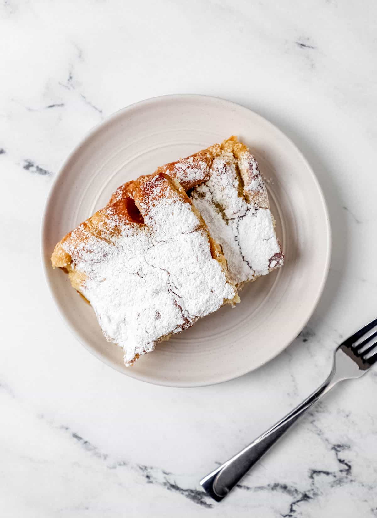 Close-up shot of a single serving of French toast casserole on a white plate with fresh berries and powdered sugar, fork cutting through showing the custardy interior texture