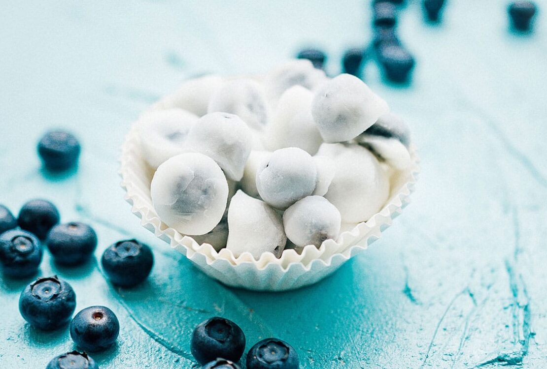 A beautiful overhead shot of frozen yogurt covered blueberries arranged on a white marble surface, glistening with a creamy white yogurt coating, scattered with fresh blueberries and a sprig of mint