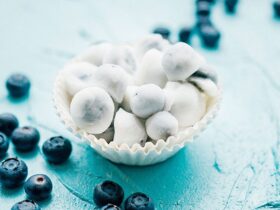 A beautiful overhead shot of frozen yogurt covered blueberries arranged on a white marble surface, glistening with a creamy white yogurt coating, scattered with fresh blueberries and a sprig of mint