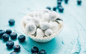 A beautiful overhead shot of frozen yogurt covered blueberries arranged on a white marble surface, glistening with a creamy white yogurt coating, scattered with fresh blueberries and a sprig of mint