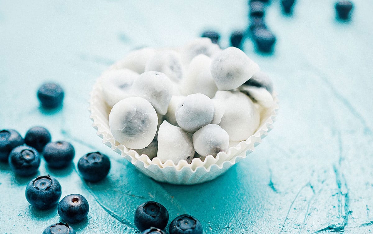 A beautiful overhead shot of frozen yogurt covered blueberries arranged on a white marble surface, glistening with a creamy white yogurt coating, scattered with fresh blueberries and a sprig of mint