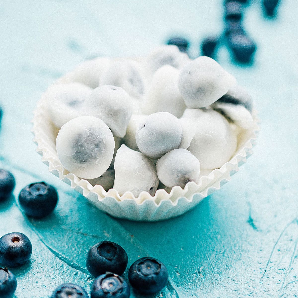 A close-up macro shot of frozen yogurt covered blueberries in a small white bowl, the yogurt coating slightly frosty and creamy, a few berries broken open to show the juicy purple interior