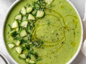 A beautifully plated bowl of Gazpacho Verde with avocado, garnished with a drizzle of olive oil, thin cucumber slices, and fresh herbs, photographed on a light marble surface with a linen napkin