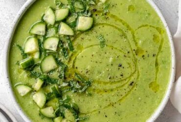 A beautifully plated bowl of Gazpacho Verde with avocado, garnished with a drizzle of olive oil, thin cucumber slices, and fresh herbs, photographed on a light marble surface with a linen napkin