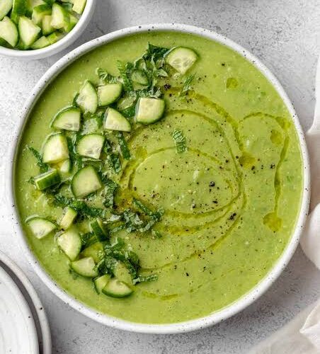A beautifully plated bowl of Gazpacho Verde with avocado, garnished with a drizzle of olive oil, thin cucumber slices, and fresh herbs, photographed on a light marble surface with a linen napkin