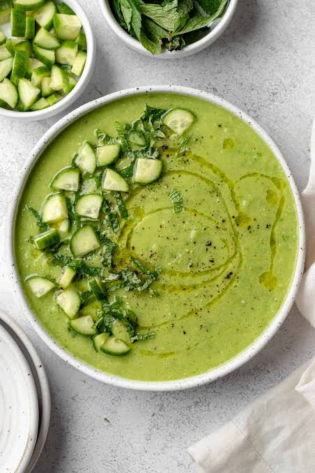 A beautifully plated bowl of Gazpacho Verde with avocado, garnished with a drizzle of olive oil, thin cucumber slices, and fresh herbs, photographed on a light marble surface with a linen napkin