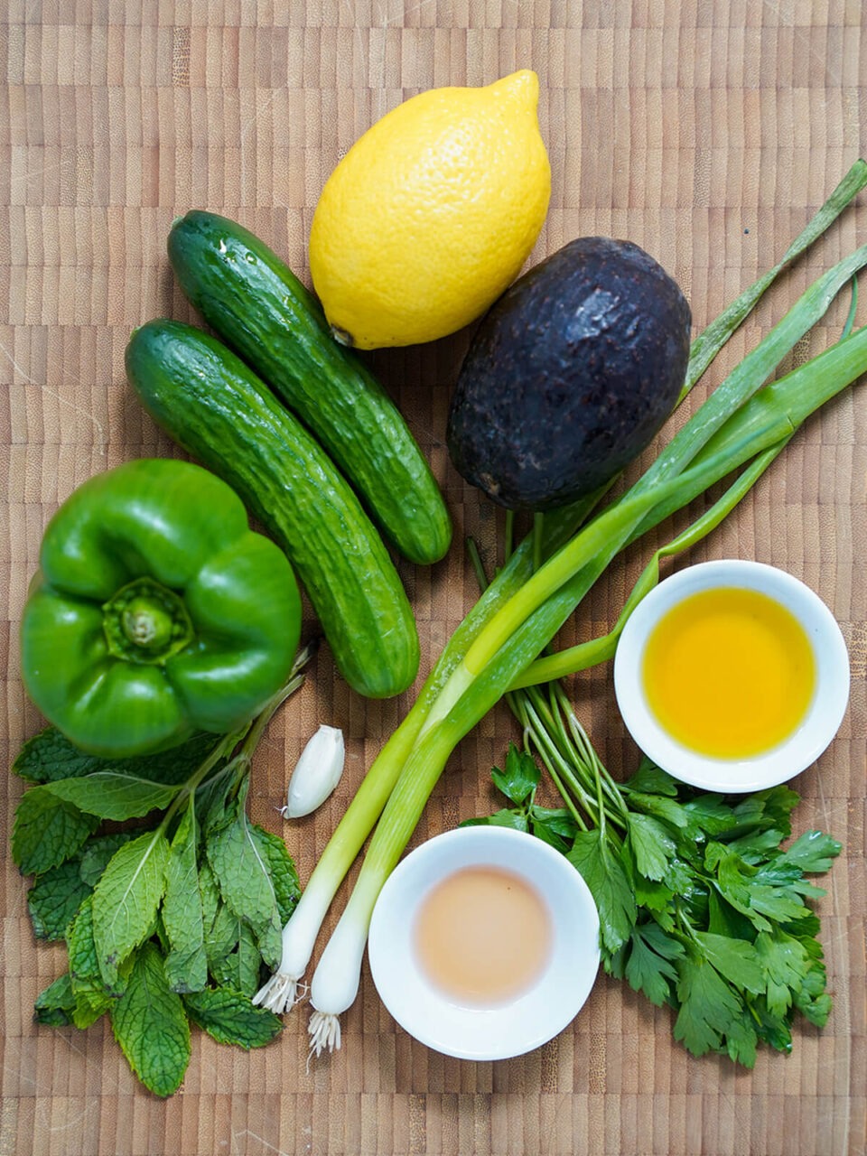 Overhead flat-lay shot of all the Gazpacho Verde ingredients laid out on a wooden cutting board — halved cucumber, avocado, green bell pepper, garlic cloves, fresh herbs, olive oil, and a lemon