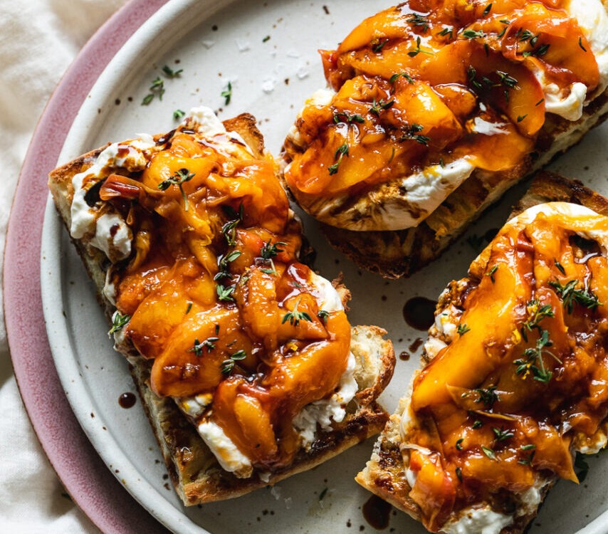 Grilled peach burrata breakfast toast on rustic wooden board with golden caramelized peaches and torn creamy burrata on thick whole grain bread slices
