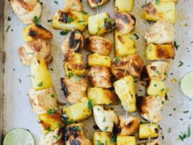 Grilled pineapple chicken skewers served on a wooden board with fresh pineapple slices, colorful bell peppers, and a drizzle of teriyaki glaze, garnished with chopped cilantro and lime wedges