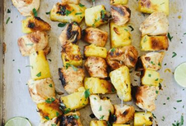 Grilled pineapple chicken skewers served on a wooden board with fresh pineapple slices, colorful bell peppers, and a drizzle of teriyaki glaze, garnished with chopped cilantro and lime wedges