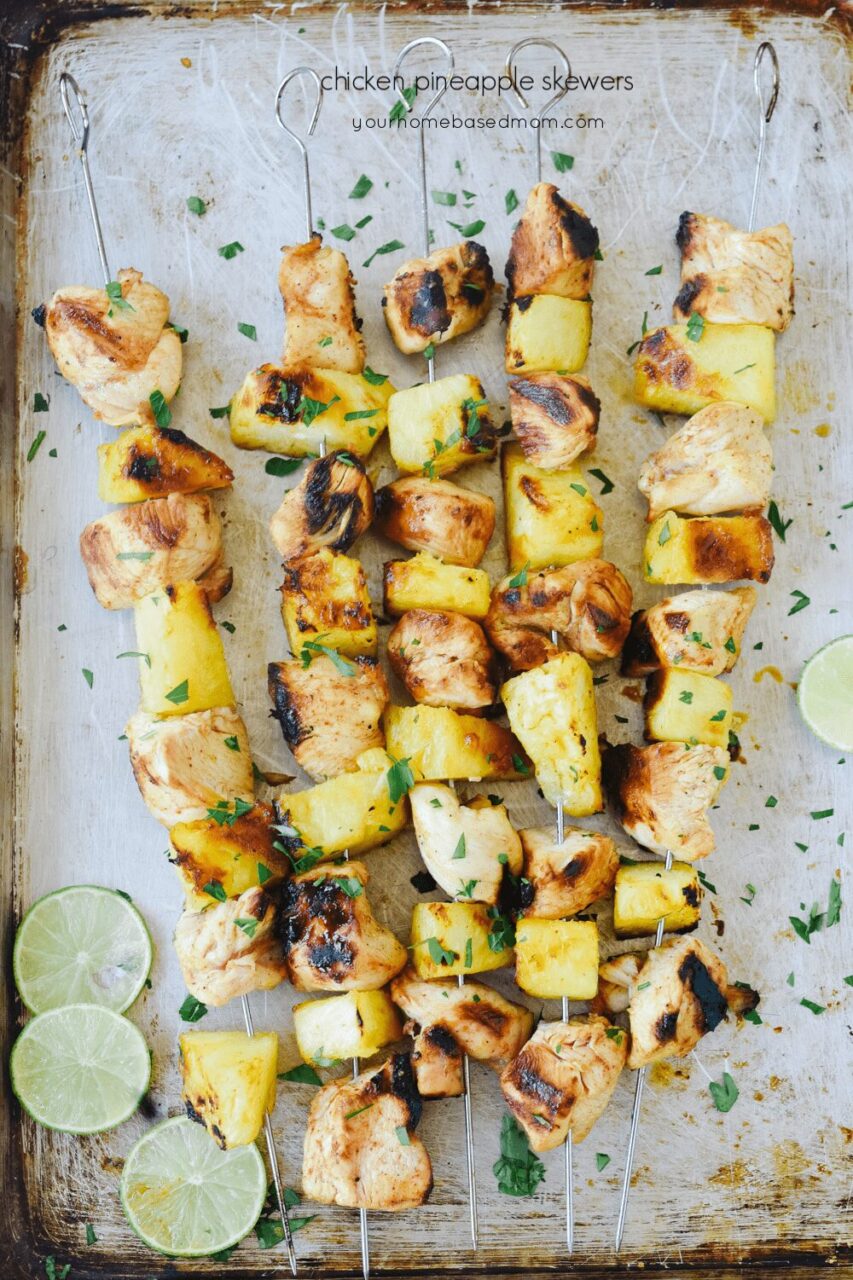 Grilled pineapple chicken skewers served on a wooden board with fresh pineapple slices, colorful bell peppers, and a drizzle of teriyaki glaze, garnished with chopped cilantro and lime wedges
