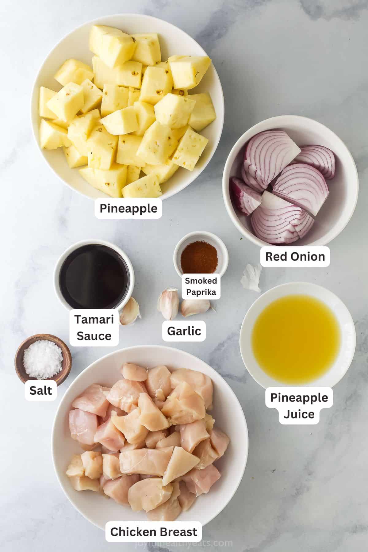 Flat lay of grilled pineapple chicken skewer ingredients including chicken thigh pieces, pineapple chunks, bell peppers, garlic cloves, soy sauce bottle, and marinade in a glass bowl on a wooden surface