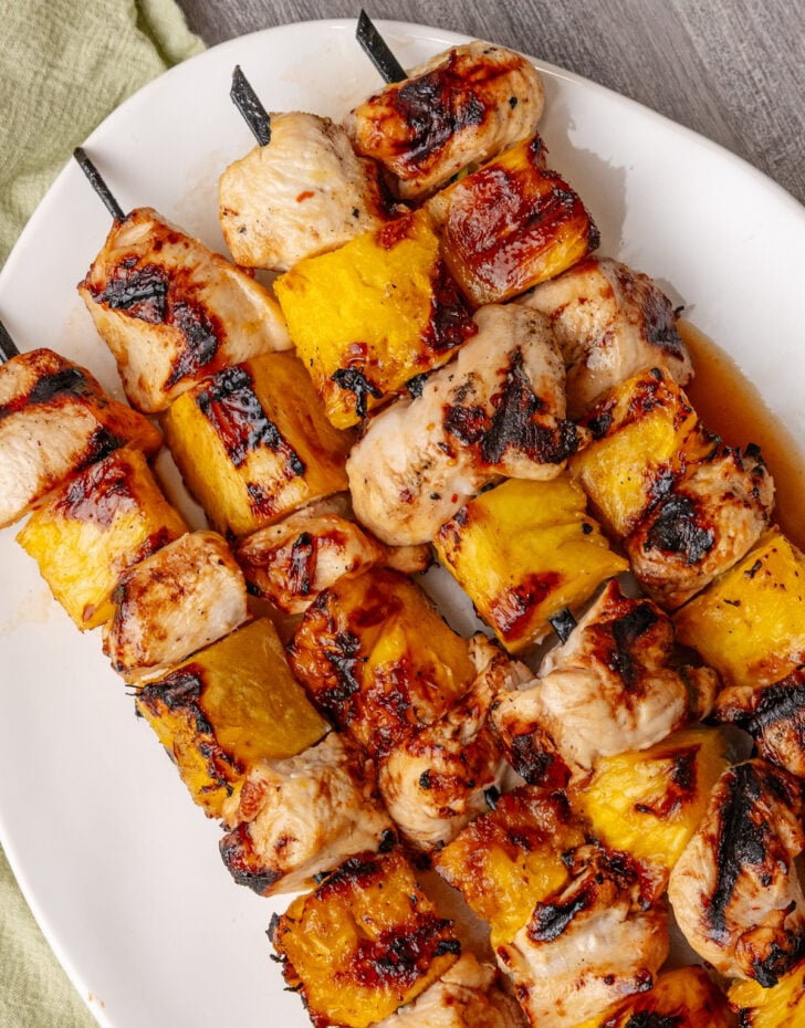 Close-up of grilled pineapple chicken skewer pieces on a plate showing caramelized char marks on juicy chicken and golden pineapple chunks, with a lime wedge and fresh herbs in the background