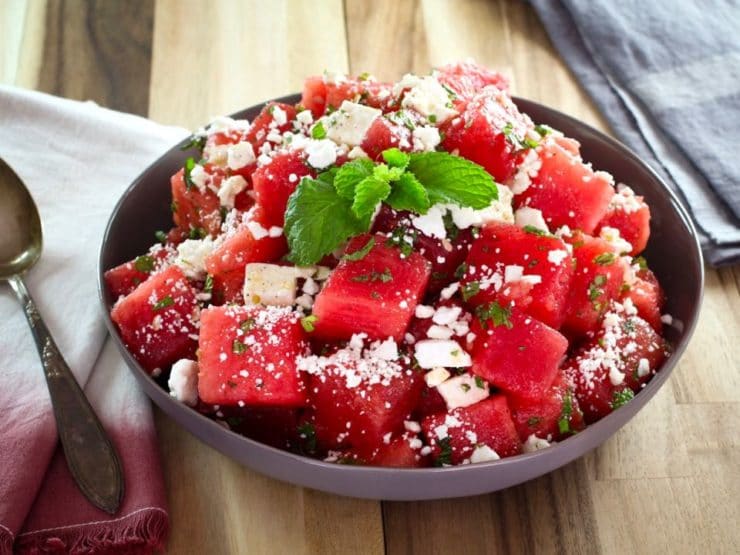 Fresh watermelon cubes, crumbled feta cheese, and fresh mint leaves arranged on a cutting board with lime halves