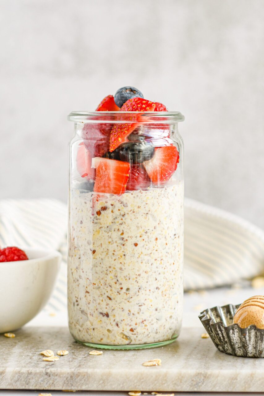 Overnight oats with summer berries in a glass mason jar, layered with Greek yogurt, strawberries, blueberries, and raspberries, topped with honey drizzle, photographed on a bright white marble surface with fresh mint garnish