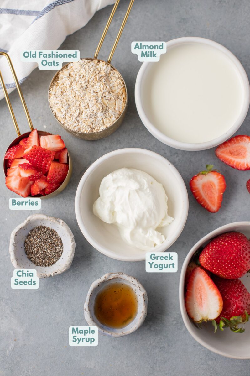 Overhead flat lay of overnight oats ingredients on a wooden cutting board: mason jars, rolled oats in a bowl, fresh strawberries and blueberries, Greek yogurt in a white bowl, chia seeds, honey jar, and almond milk carton