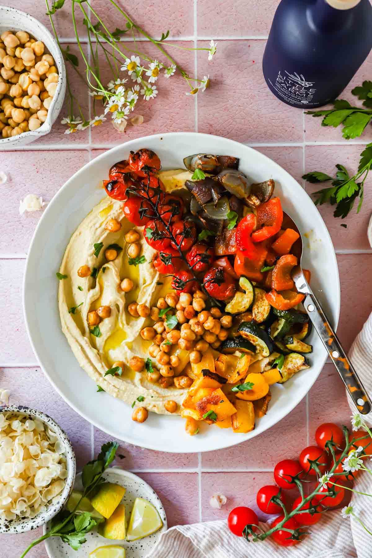Overhead flat-lay of hummus bowl ingredients including chickpeas, tahini, spices, colorful vegetables, olive oil, and fresh herbs on a wooden surface
